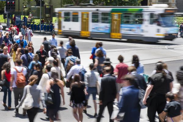 A blurred out tram as a crowd of people cross a road in the foreground
